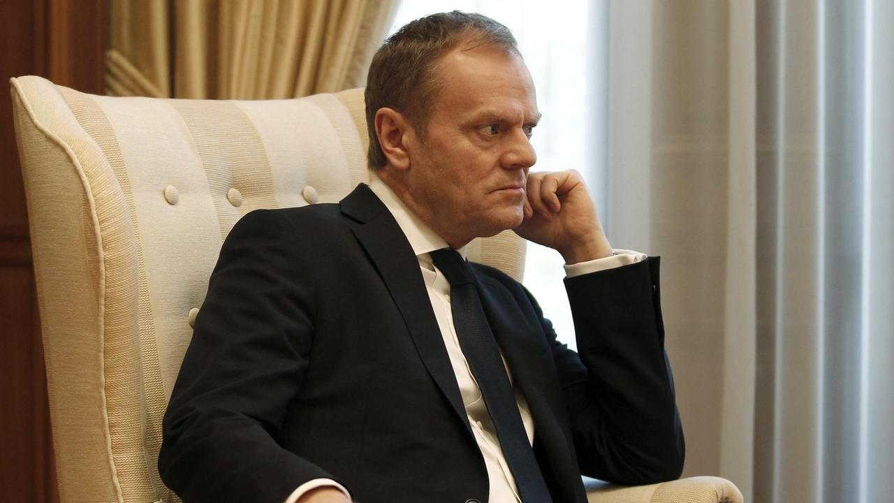 Tusk sitzt in einem beigen Sessel vor einem Fenster mit weißem Vorhang und hat eine Hand an das Kinn gestützt. Er blickt konzentriert und scheint jemandem aufmerksam zuzuhören. Tusk sitzt in einem beigen Sessel vor einem Fenster mit weißem Vorhang und hat eine Hand an das Kinn gestützt. Er blickt konzentriert und scheint jemandem aufmerksam zuzuhören.