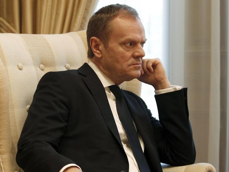 Tusk sitzt in einem beigen Sessel vor einem Fenster mit weißem Vorhang und hat eine Hand an das Kinn gestützt. Er blickt konzentriert und scheint jemandem aufmerksam zuzuhören.