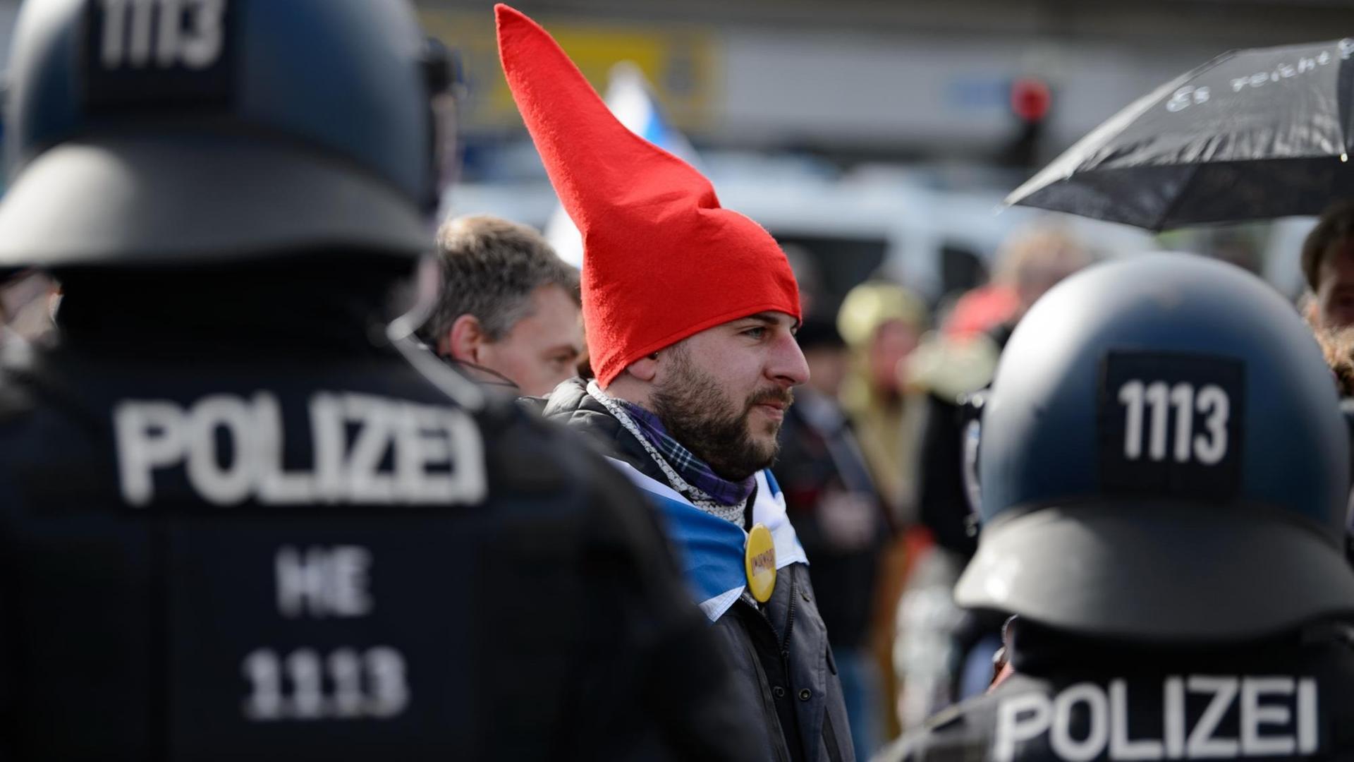 Ein Teilnehmer mit einer roten Zipfelmütze und ohen Mund-Nasen-Bedeckung steht bei einer Kundgebung unter dem Motto "Freie Bürger Kassel - Grundrechte und Demokratie" vor Polizisten. Ein Teilnehmer mit einer roten Zipfelmütze und ohen Mund-Nasen-Bedeckung steht bei einer Kundgebung unter dem Motto "Freie Bürger Kassel - Grundrechte und Demokratie" vor Polizisten.