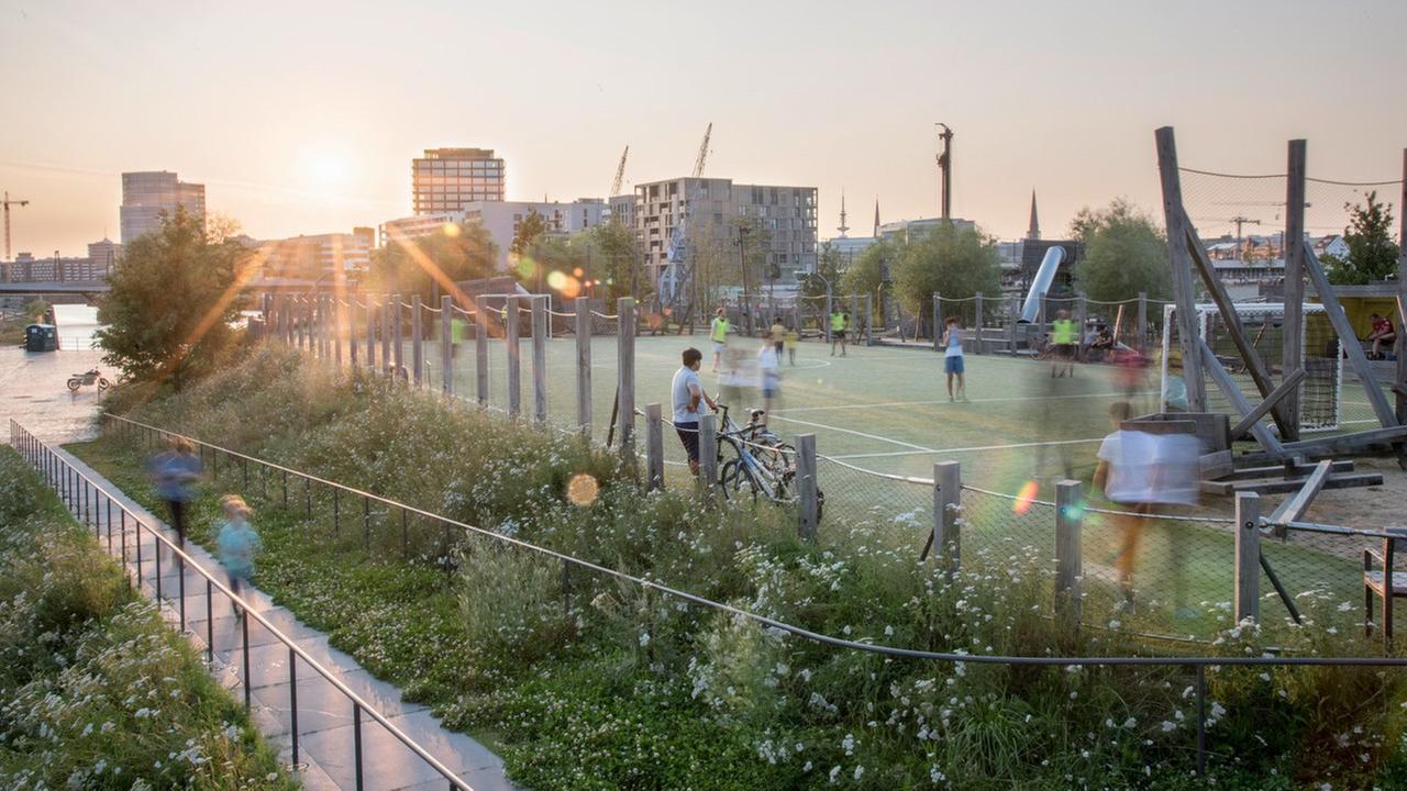 Der Baakenpark in Hamburg, entworfen vom Atelier Loidl. Zu sehen ist ein kleiner Fußballplatz, auf dem Menschen Fußball spielen. Drum herum wachsen Pflanzen. Im Hintergrund sieht man moderne Wohnhäuser. Der Baakenpark in Hamburg, entworfen vom Atelier Loidl. Zu sehen ist ein kleiner Fußballplatz, auf dem Menschen Fußball spielen. Drum herum wachsen Pflanzen. Im Hintergrund sieht man moderne Wohnhäuser.