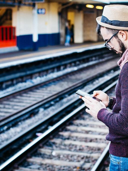 Ein junger Mann steht auf einem Bahnsteig und liest auf seinem Smartphone