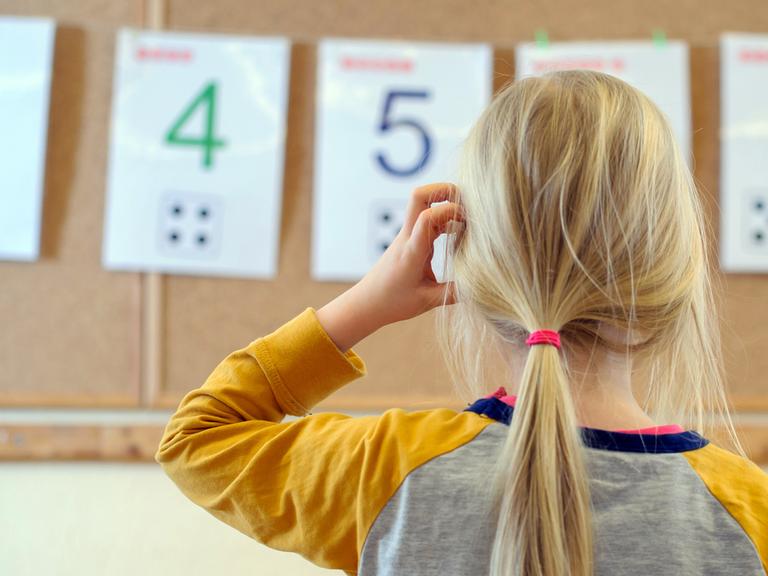 Ein Schulkind steht nachdenklich vor einer an der Wand aufgehängten Zahlenreihe.