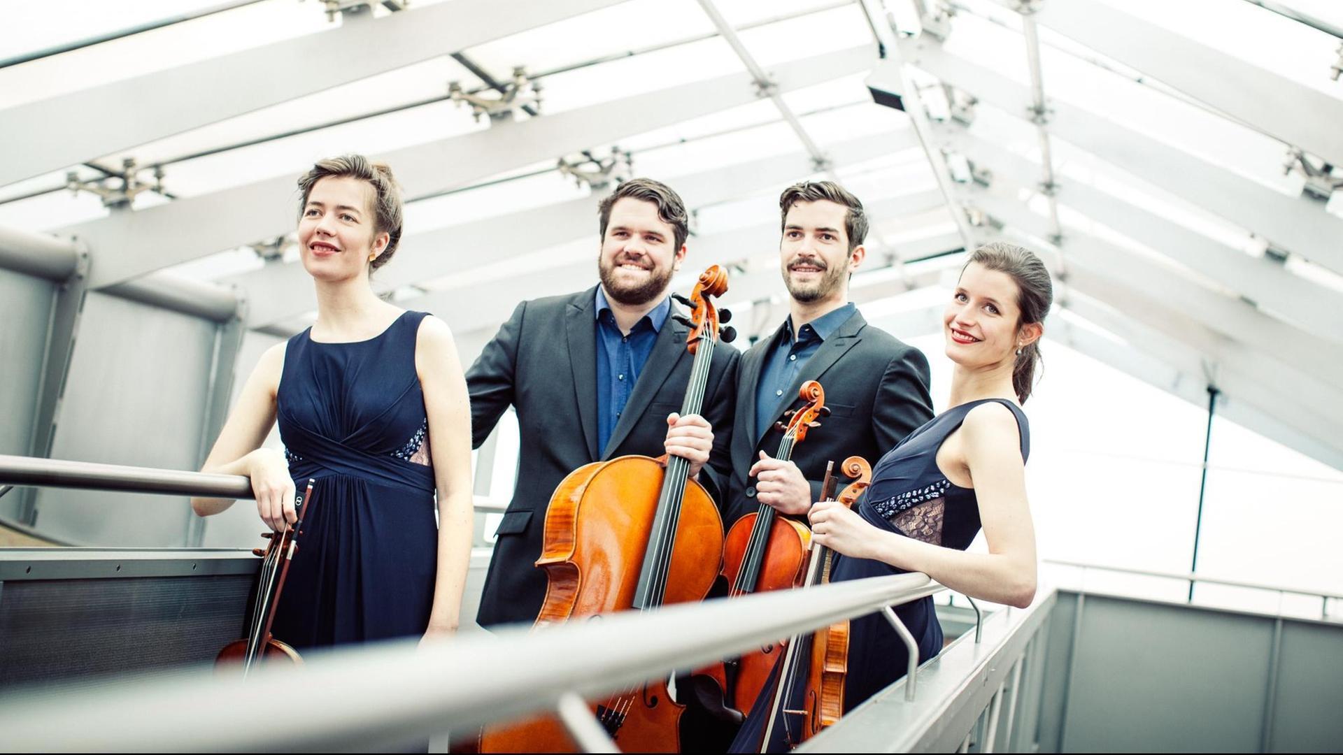 Zwei Musikerinnen und zwei Musiker stehen in einer luftigen, modernen Glashalle. Sie tragen blaue Kleider beziehungsweise Hemden und halten ihre Streichinstrumente in der Hand. Zwei Musikerinnen und zwei Musiker stehen in einer luftigen, modernen Glashalle. Sie tragen blaue Kleider beziehungsweise Hemden und halten ihre Streichinstrumente in der Hand.
