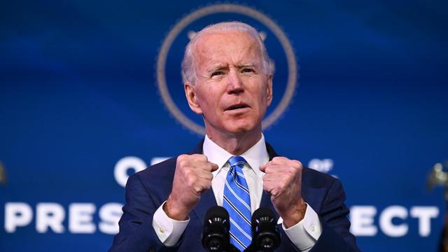 Joe Biden hat während einer Rede auf einem Podium beide Hände erhoben.
