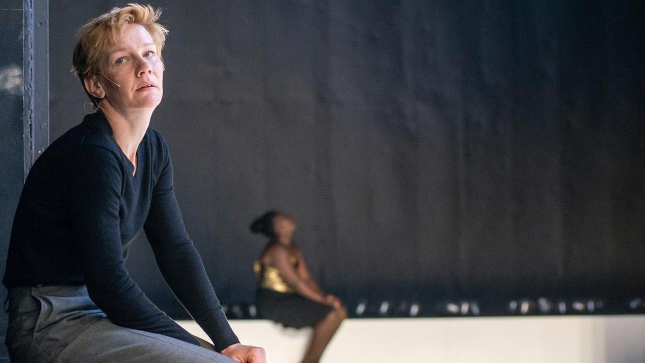 Sandra Hüller in der Rolle von Hamlet sitzt auf einem Podest und blickt seitlich in die Kamera. Im Hintergrund ist Mercy Dorcas Otieno zu sehen. Sandra Hüller in der Rolle von Hamlet sitzt auf einem Podest und blickt seitlich in die Kamera. Im Hintergrund ist Mercy Dorcas Otieno zu sehen.