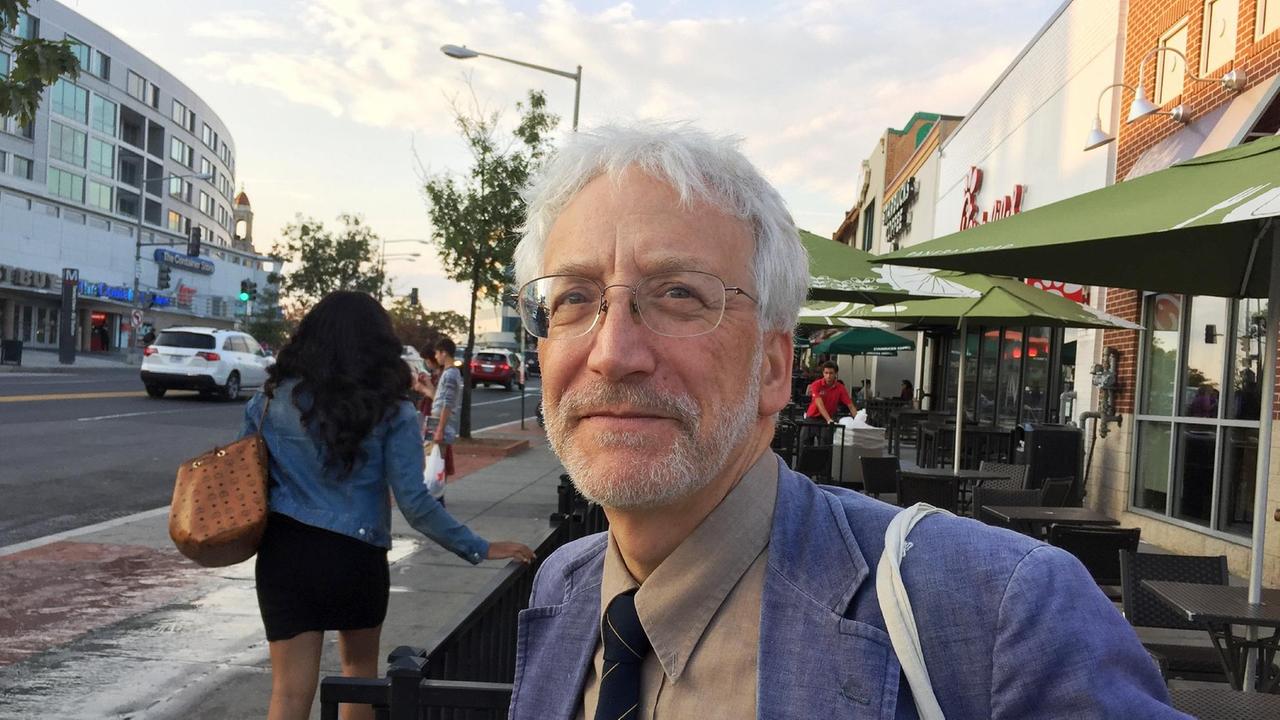 Ein Mann mit weißen Haaren, grauem Bart und dezenter Brille steht in blauem Sakko an einer Straße. Ein Mann mit weißen Haaren, grauem Bart und dezenter Brille steht in blauem Sakko an einer Straße.