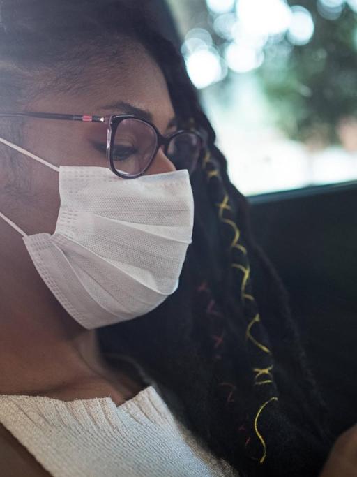 Frau sitzt auf dem Rücksitz eines Autos mit Maske und schaut auf ihr Smartphone