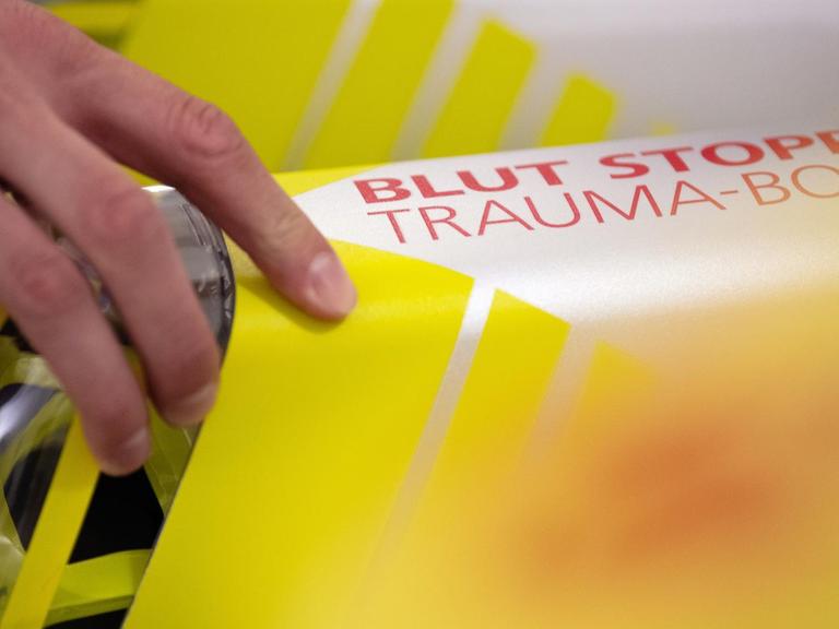 Eine Hand greift nach der Trauma-Box mit der Aufschrift «Blut Stopp». Die Box enthält ein sogenanntes Tourniquet, ein Abbindesystem ähnlich einer Blutdruckmanschette, und einen saugfähigen Druckverband zum Stillen einer Blutung eines Verletzten, bis medizinisches Fachpersonal eintrifft.