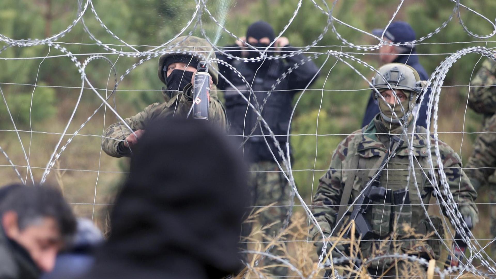 Polnische Grenzpolizisten stehen an einem Stacheldrahtzaun, hinter dem Migranten stehen.