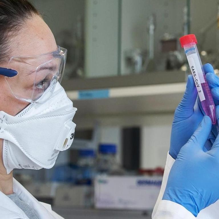 Eine Wissenschaftlerin mit Mundschutz, Schutzbrille und Handschuhen hält ein Laborröhrchen mit der Aufschrift "SARS-CoV 2" in den Händen.