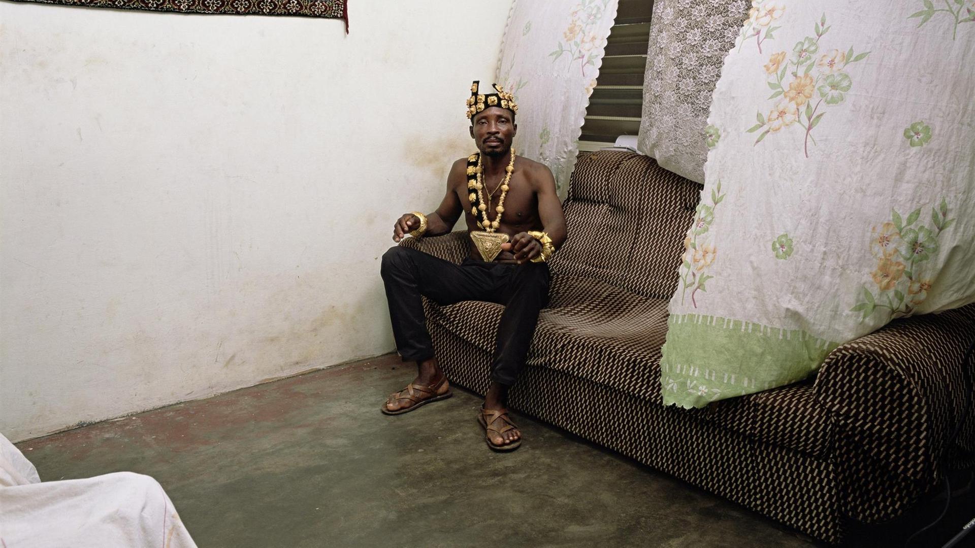 Deanna Lawson: Chief, 2019. Das Foto zeigt einen Mann mit Goldkrone und Goldkette auf einem alten Sofa sitzend. Deanna Lawson: Chief, 2019. Das Foto zeigt einen Mann mit Goldkrone und Goldkette auf einem alten Sofa sitzend.