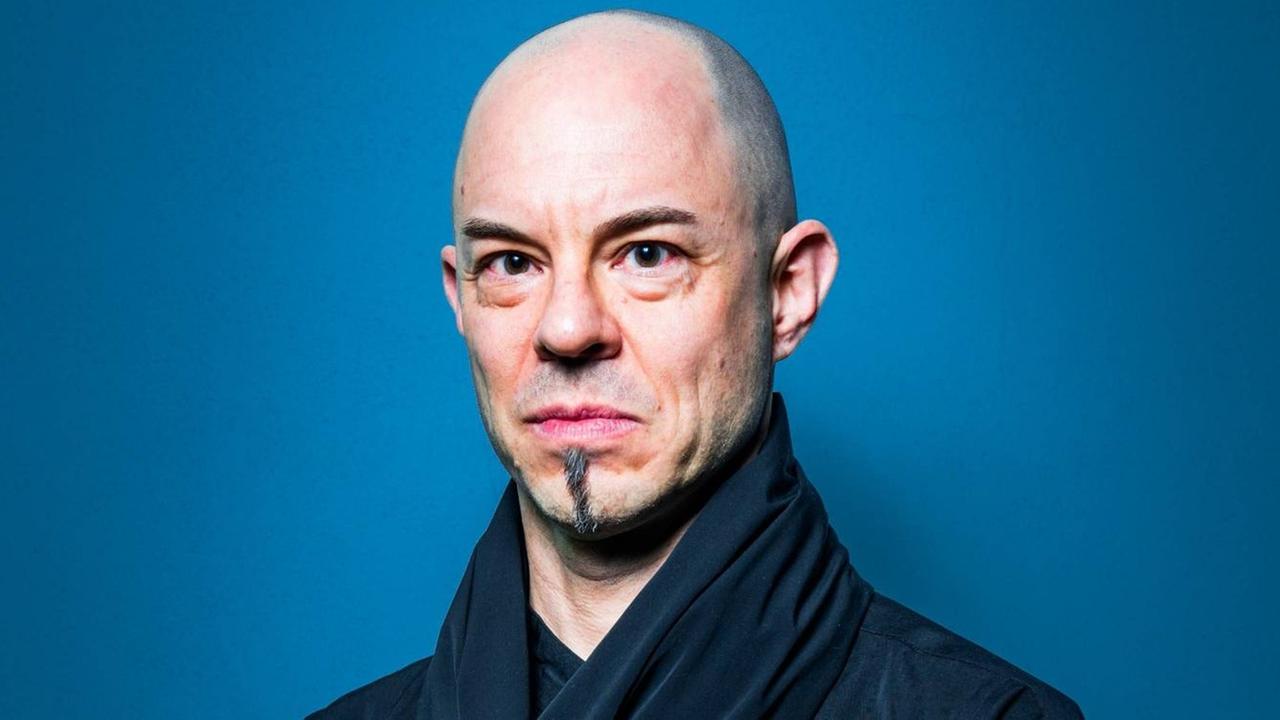 Ein glatzköpfiger Mann steht vor einem blauen Hintergrund und blickt ernst in die Kamera. Ein glatzköpfiger Mann steht vor einem blauen Hintergrund und blickt ernst in die Kamera.