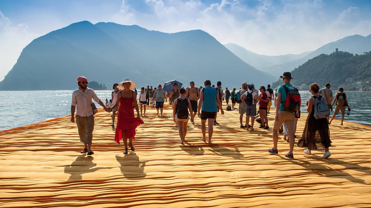 Menschen laufen bei Sonnenschein auf der Installation "Floating Piers" des bulgarischen Künstlers Christo im Sommer 2016 auf dem Iseo-See - im Hintergrund sind Berge zu sehen. Menschen laufen bei Sonnenschein auf der Installation "Floating Piers" des bulgarischen Künstlers Christo im Sommer 2016 auf dem Iseo-See - im Hintergrund sind Berge zu sehen.