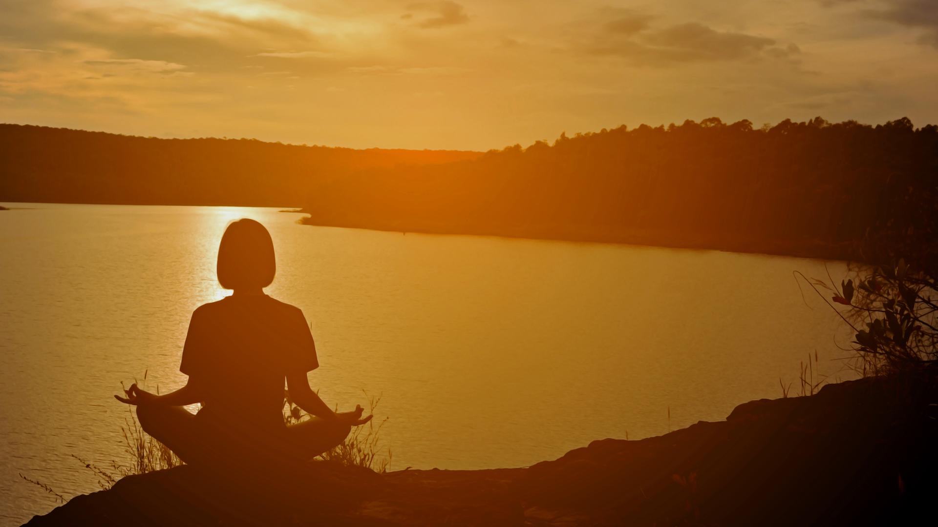 Eine Frau in Meditationshaltung sitzt in der Abendsonne auf einem Hügel mit Blick auf einen See. Eine Frau in Meditationshaltung sitzt in der Abendsonne auf einem Hügel mit Blick auf einen See.
