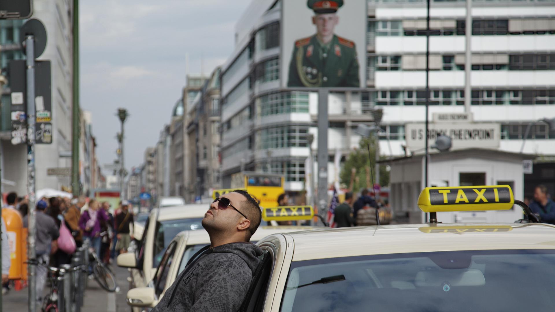 Ein Taxifahrer hält einen Moment inne und lehnt sich gegen sein Taxi am Checkpoint Charlie in Berlin. Ein Taxifahrer hält einen Moment inne und lehnt sich gegen sein Taxi am Checkpoint Charlie in Berlin.