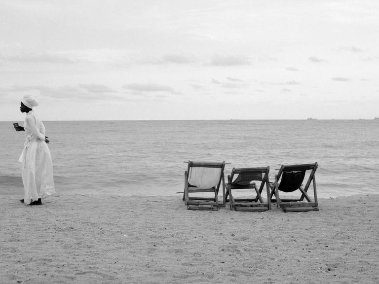 Eine Frau in weißem Kleid steht an einem Strand, neben ihr sind drei Standliegen.