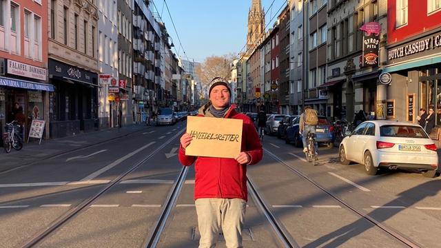 Ein Mann steht auf einer leeren Straße in Köln und hält ein Pappschild mit der Aufschrift "Veedelsretter"