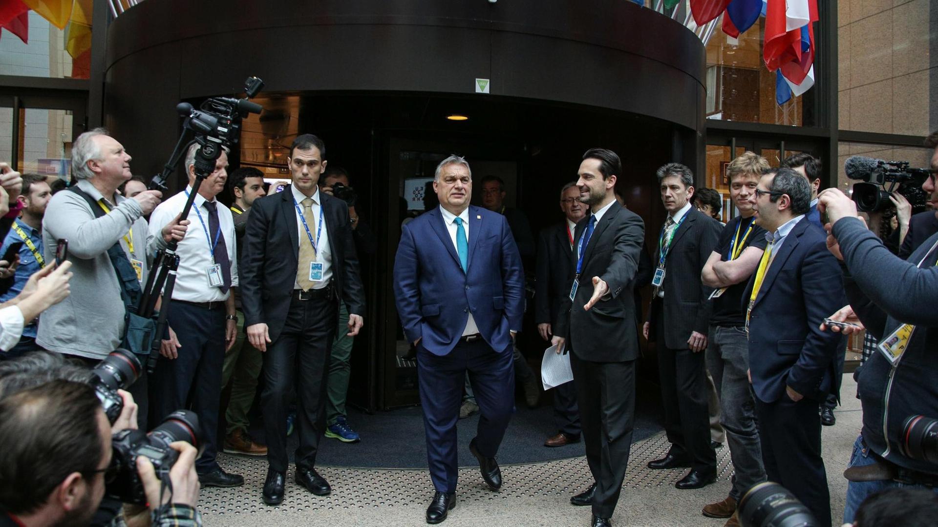 Ungarns Premierminister Victor Orbán verlässt nach einem Treffen des Europarates mit den Händen in den Hosentaschen ein Gebäude in Brüssel mit Fahnen europäischer Staaten über dem Eingang. Ungarns Premierminister Victor Orbán verlässt nach einem Treffen des Europarates mit den Händen in den Hosentaschen ein Gebäude in Brüssel mit Fahnen europäischer Staaten über dem Eingang.