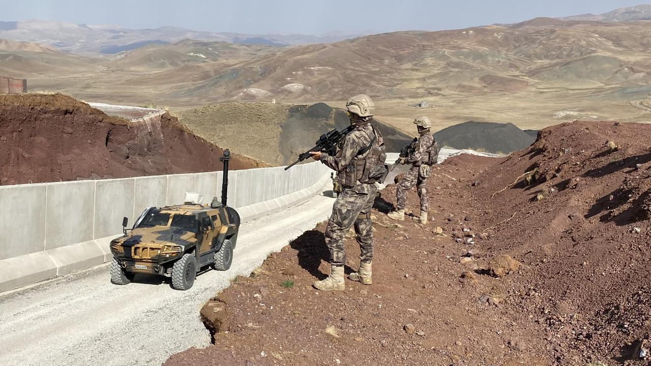 Ein Militärfahrzeug fährt eine Mauer entlang, von oben von bewaffneten Männern beobachtet. Ein Militärfahrzeug fährt eine Mauer entlang, von oben von bewaffneten Männern beobachtet.