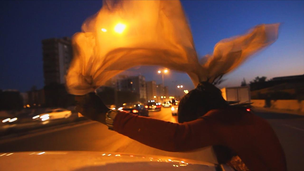 Szene aus dem Film "Freedom Fields": In der Abenddämmerung schwenkt eine weibliche Silhouette aus einem fahrenden Auto ein gelb leuchtendes Tuch.
