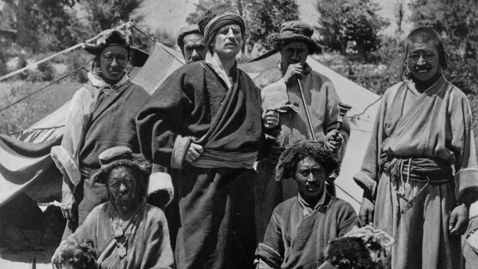 Schwarz-weiß-Aufnahme von einer Gruppe Tibetaner, in deren Mitte Sven Hedin in deren traditioneller Kleidung steht. Schwarz-weiß-Aufnahme von einer Gruppe Tibetaner, in deren Mitte Sven Hedin in deren traditioneller Kleidung steht.