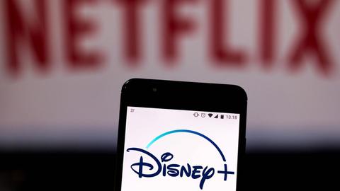 Auf einem Handy leuchtet das Logo von Disney+, im Hintergrund verschwommen das Netflix-Logo.