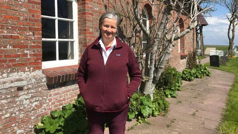 Pfarrerin Hildegard Rugenstein steht in einer weinroten Outdoor-Jacke vor ihrer Kirche: einem Haus mit roter Ziegelmauer und einem reetgedeckten Dach. Pfarrerin Hildegard Rugenstein steht in einer weinroten Outdoor-Jacke vor ihrer Kirche: einem Haus mit roter Ziegelmauer und einem reetgedeckten Dach.