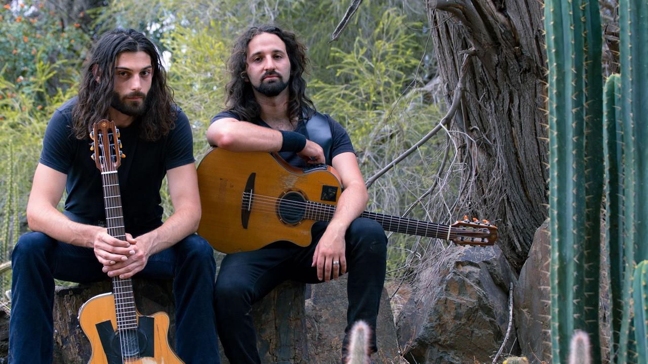 Zwei Männer mit langen, schwarzen Haaren und akustischen Gitarren sitzen auf einem Stein nebeneinander.