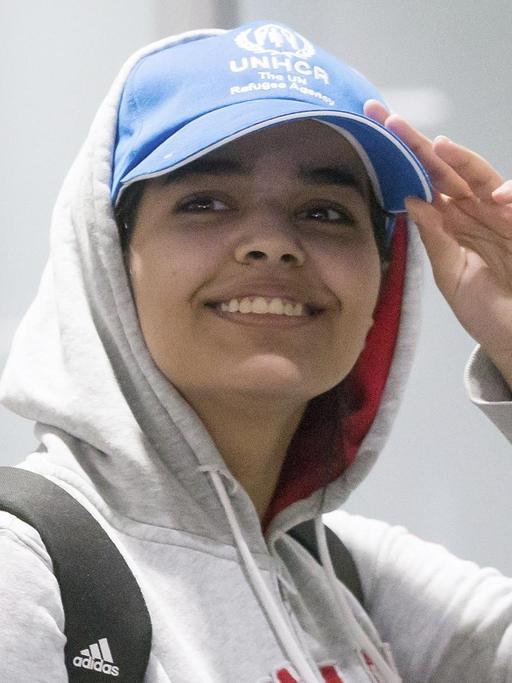 Die junge Frau trägt ein graues Sweat-Shirt mit der Aufschrift "Kanada" und eine blaue Baseball-Cap des UNHCR. Sie lächelt breit und greift an den Schirm ihrer Mütze.