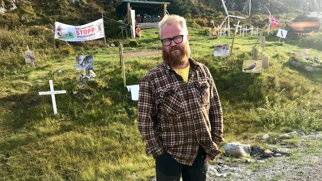 Ein stämmiger Mann in dunkelbrauner Kleidung mit buschigem roten Vollbart, blonden Haaren und mit Brille steht - die Hände in den Hosentaschen - vor diversen Protestutensilien wie weißen Kreuzen, einem Transparent und diversen Vogelplakaten. Ein stämmiger Mann in dunkelbrauner Kleidung mit buschigem roten Vollbart, blonden Haaren und mit Brille steht - die Hände in den Hosentaschen - vor diversen Protestutensilien wie weißen Kreuzen, einem Transparent und diversen Vogelplakaten.