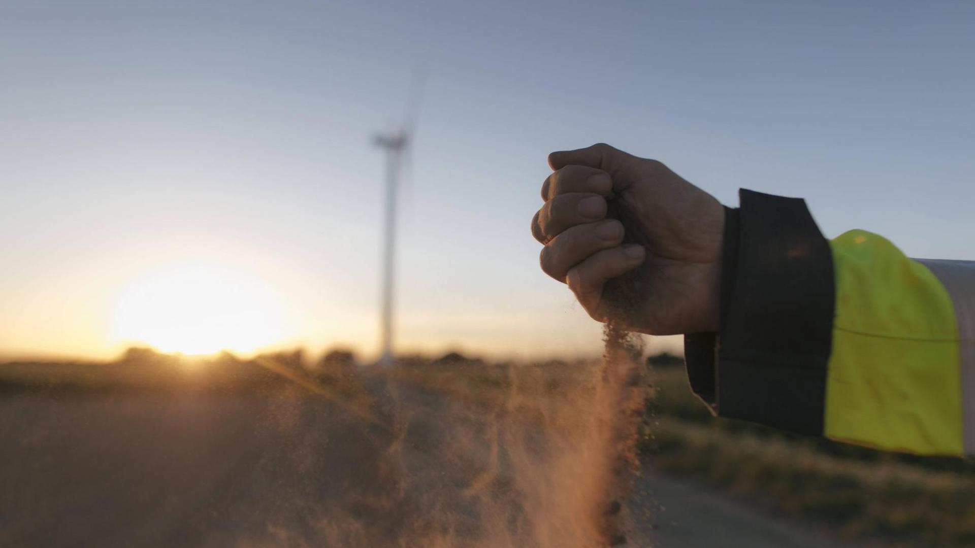 Ein Mann lässt Sand durch seine Hand rieseln, im Hintergrund ein Windrad auf einem Feld im Sonnenuntergang.