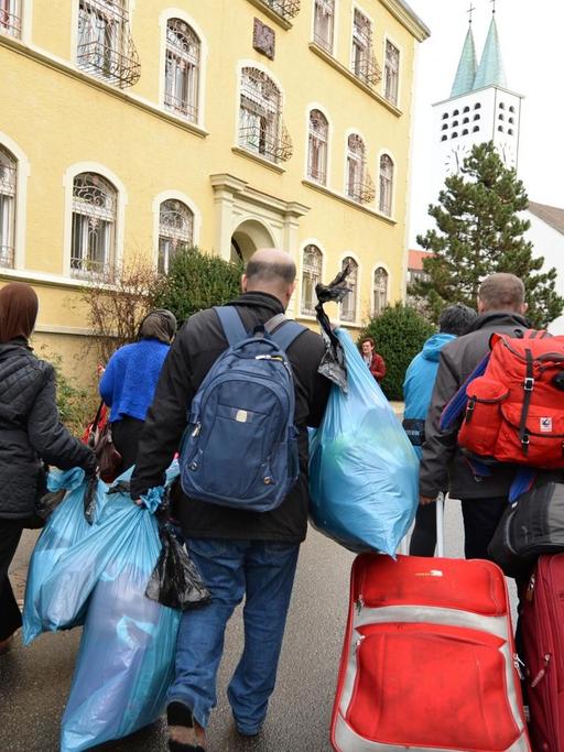 Asylbewerber aus Syrien laufen am 18.12.2015 mit ihrem Gepäck zur der Kirche St. Maria der Stiftung Liebenau in Liebenau (Baden-Württemberg). In Liebenau sollen 60 Asylbewerber zunächst für ein Jahr unterkommen.