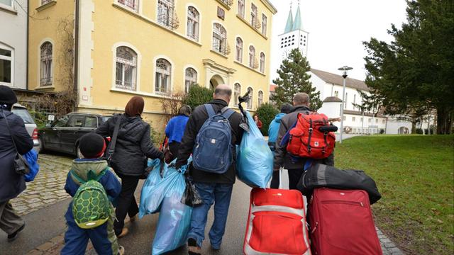 Asylbewerber aus Syrien laufen am 18.12.2015 mit ihrem Gepäck zur der Kirche St. Maria der Stiftung Liebenau in Liebenau (Baden-Württemberg). In Liebenau sollen 60 Asylbewerber zunächst für ein Jahr unterkommen.