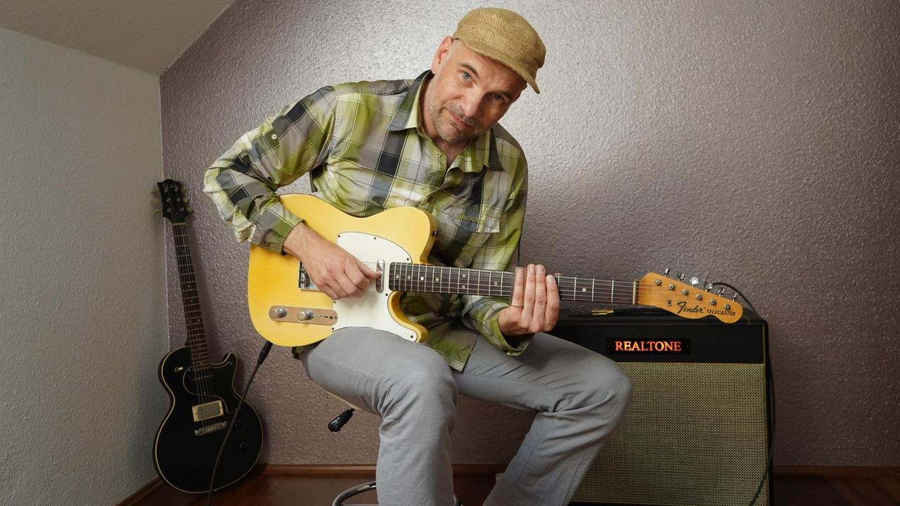 Ein Mann in kariertem Hemd sitzt mit Elektrogitarre auf einem Hocker vor dem Gitarren-Verstärker.
