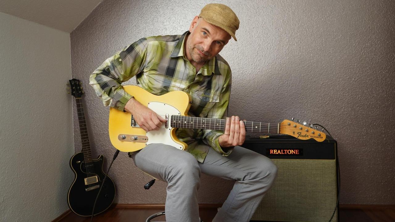 Ein Mann in kariertem Hemd sitzt mit Elektrogitarre auf einem Hocker vor dem Gitarren-Verstärker. Ein Mann in kariertem Hemd sitzt mit Elektrogitarre auf einem Hocker vor dem Gitarren-Verstärker.