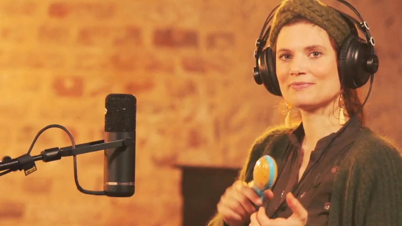 Eine Frau sitzt vor einem Mikrofon, trägt Kopfhörer und blickt in die Kamera. In der Hand hält sie einen Shaker. Eine Frau sitzt vor einem Mikrofon, trägt Kopfhörer und blickt in die Kamera. In der Hand hält sie einen Shaker.