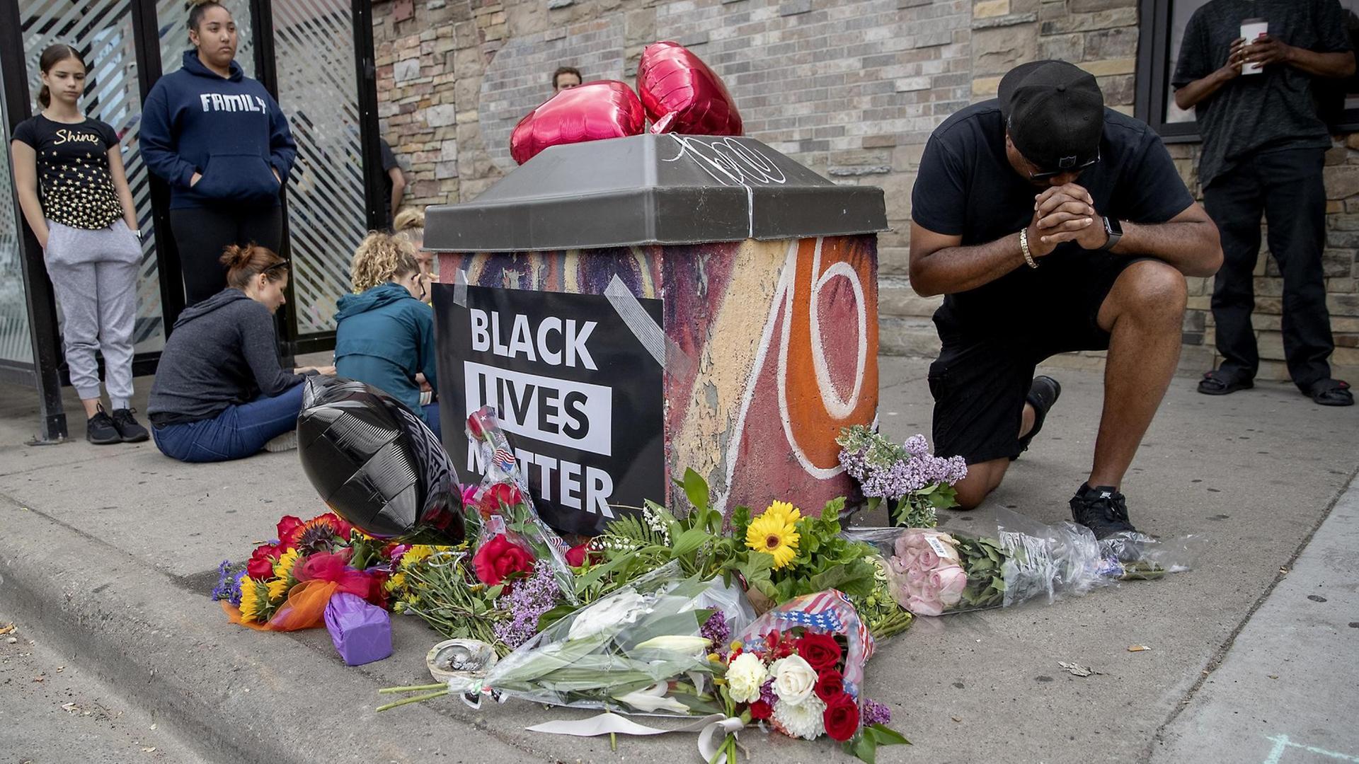 An einer Gedenkstelle mit Blumen und einem "Black Lives Matter"-Plakat trauern Menschen um George Floyd. An einer Gedenkstelle mit Blumen und einem "Black Lives Matter"-Plakat trauern Menschen um George Floyd.