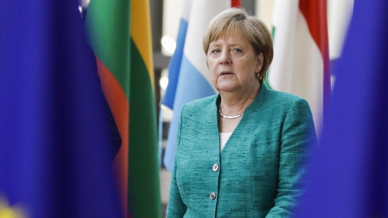 Merkel geht durch einen Gang, sie ist zwischen Flaggen der EU und nationalen Flaggen zu sehen.