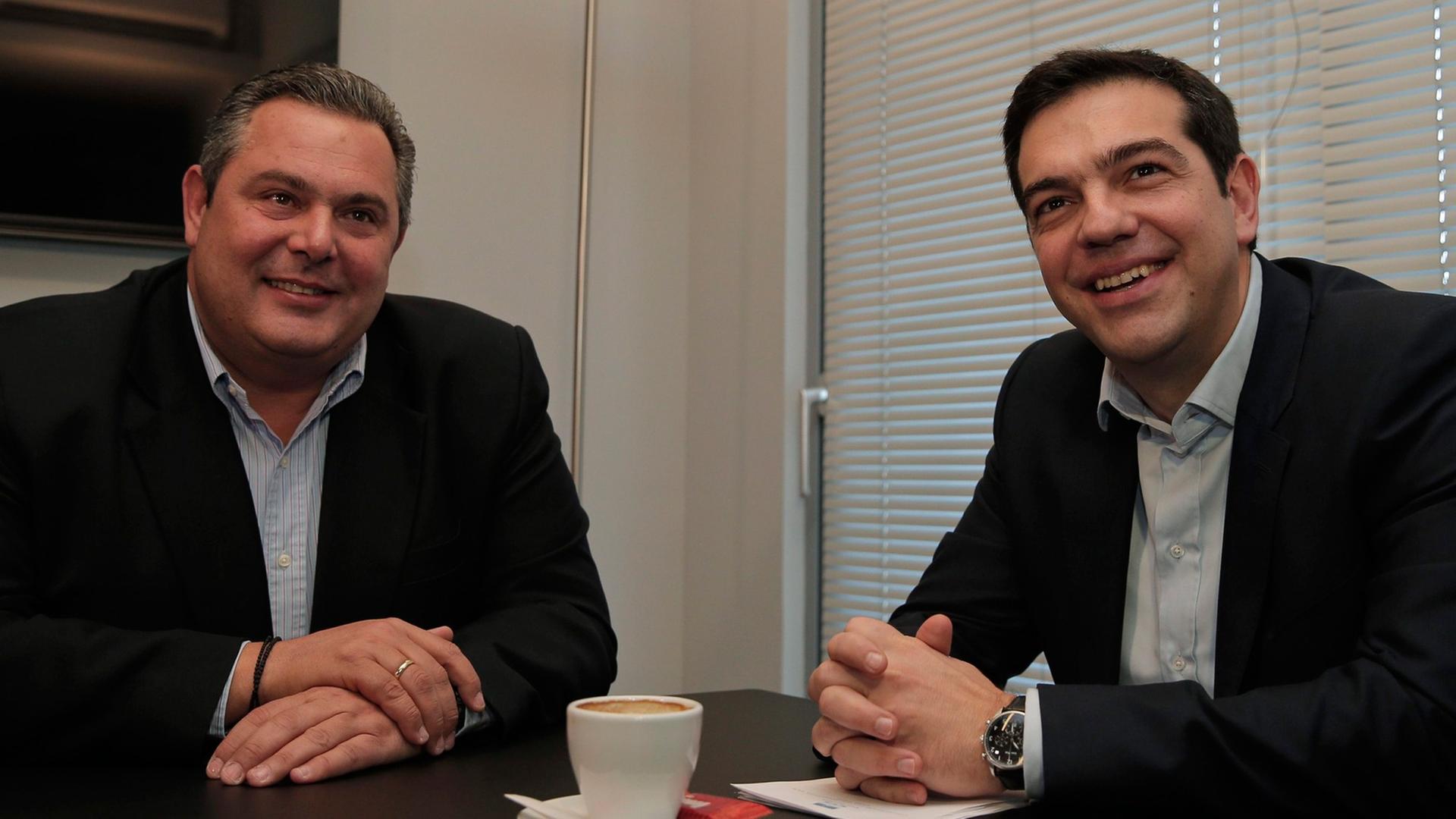 Panos Kammenos und Alexis Tsipras sitzen an einem Tisch und lächeln in die Kameras.
