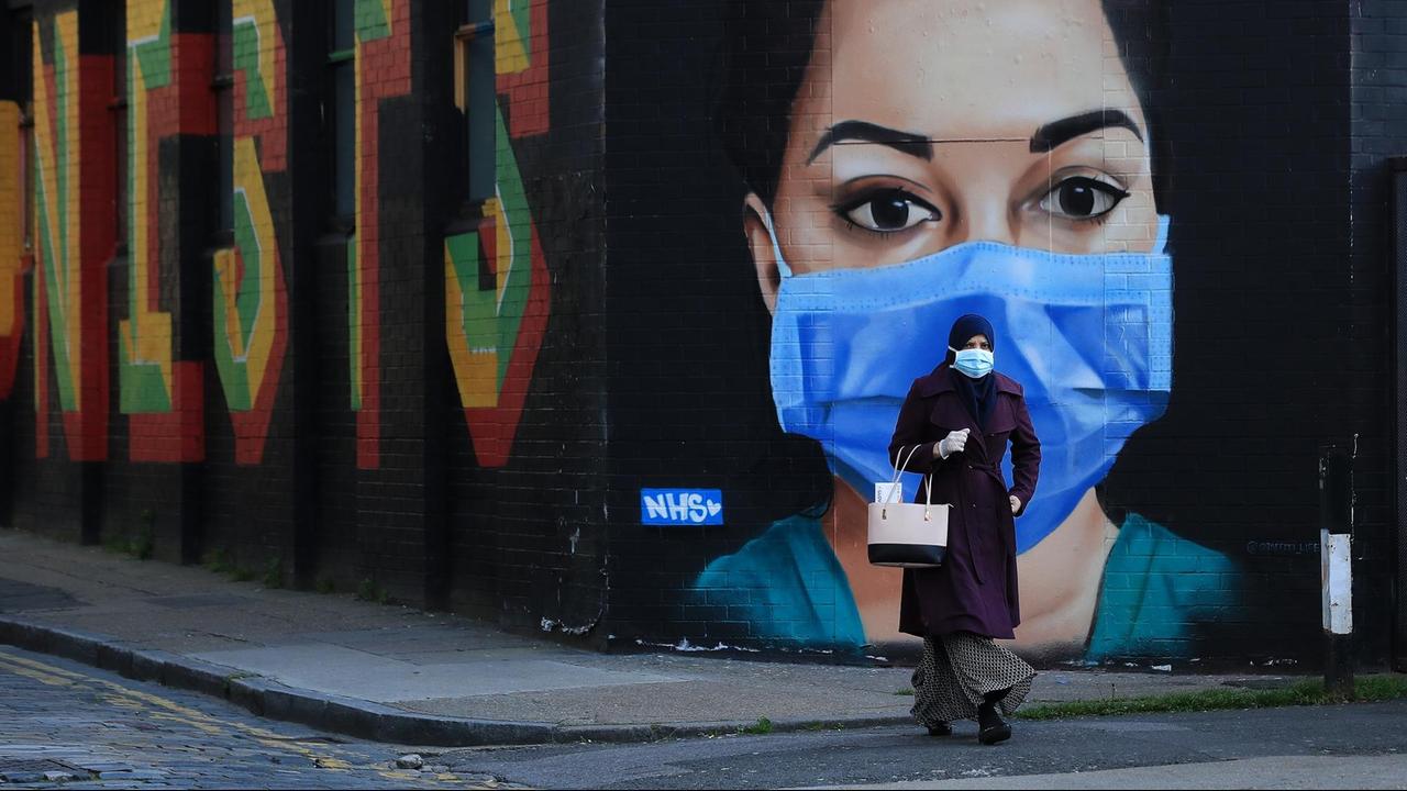 Eine Frau, die ein Kopftuch und eine Gesichtsmaske trägt, geht am 21. April 2020 im Londoner Stadtteil Shoreditch an einem Straßenkunstwerk vorbei, das einen NHS-Mitarbeiter darstellt. 