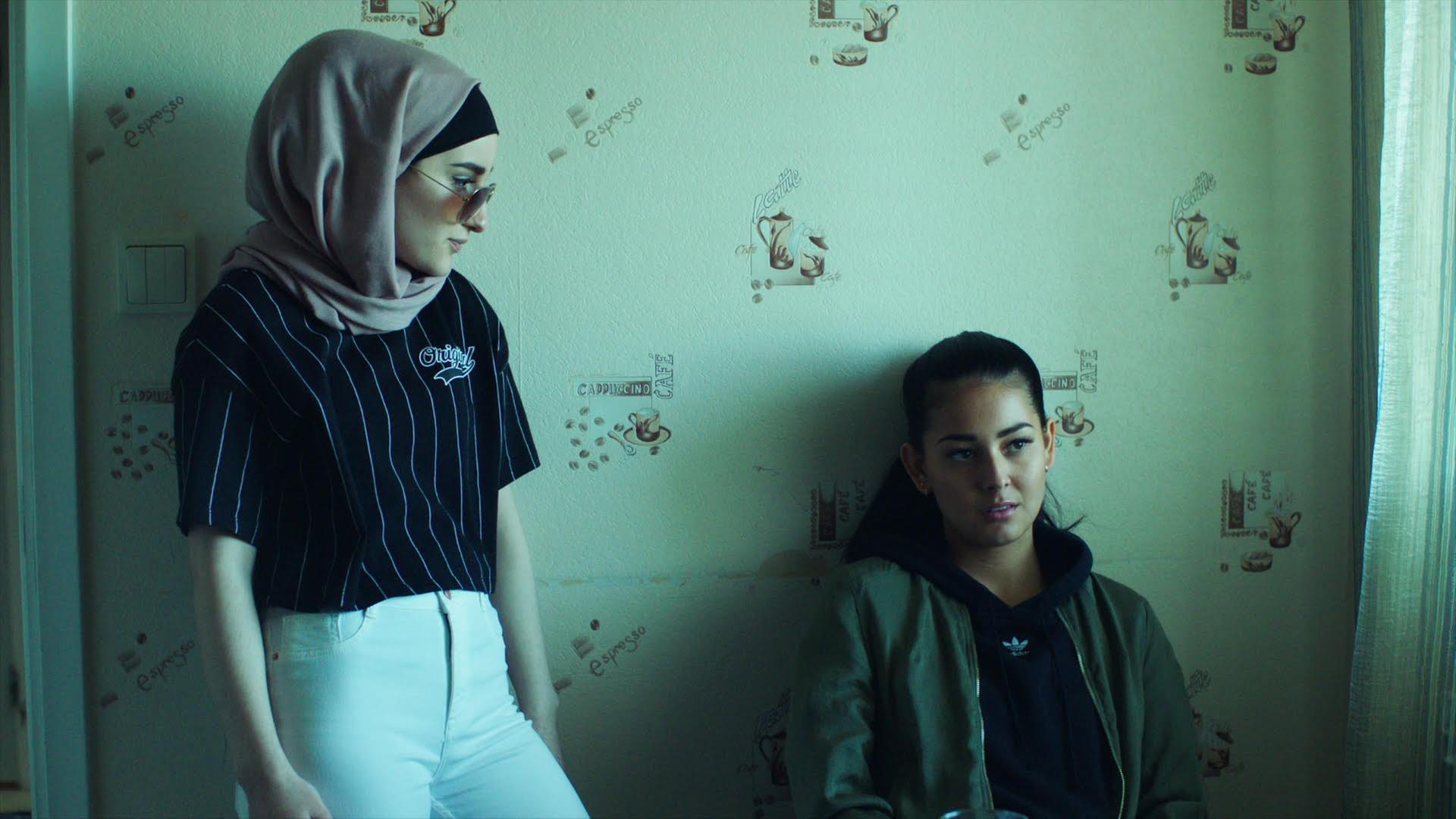 Eine Filmszene aus der Netflix-Serie Kalifat zeigt zwei Frauen. Eine trägt ein Kopftuch und steht, die zeit hockt an einer grünen Wand. Eine Filmszene aus der Netflix-Serie Kalifat zeigt zwei Frauen. Eine trägt ein Kopftuch und steht, die zeit hockt an einer grünen Wand.