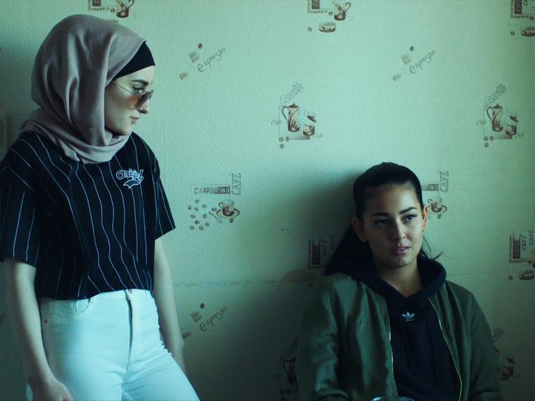 Eine Filmszene aus der Netflix-Serie Kalifat zeigt zwei Frauen. Eine trägt ein Kopftuch und steht, die zeit hockt an einer grünen Wand.