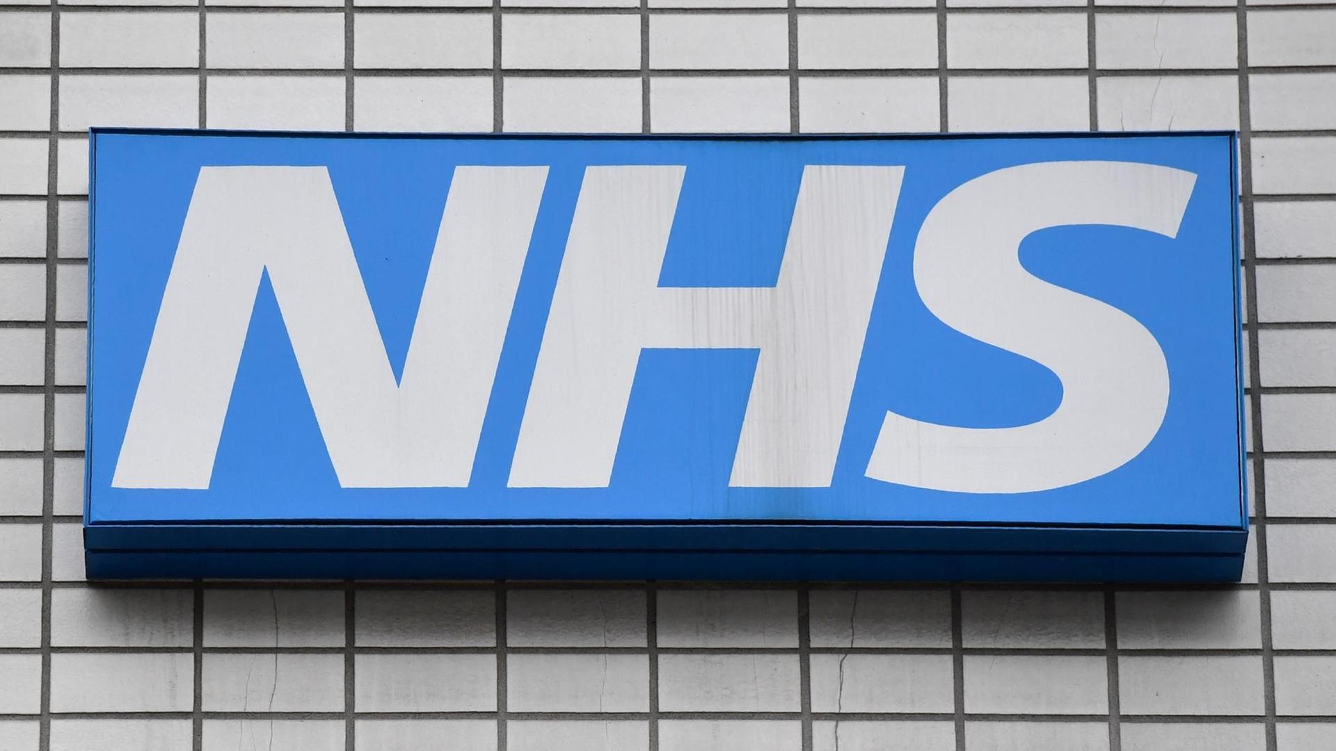 Die weißen Großbuchstaben NHS auf blauem Untergrund prangen auf einer grauen Hauswand. Die weißen Großbuchstaben NHS auf blauem Untergrund prangen auf einer grauen Hauswand.