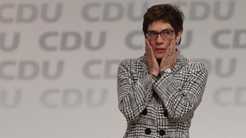 Annegret Kramp-Karrenbauer steht in einem schwarz-weiß-karrierten Sakko auf der Bühne, die Aufschrift "CDU" ist mehrfach hinter ihr zu sehen. Sie hält ihre Hände an ihre Wangen.