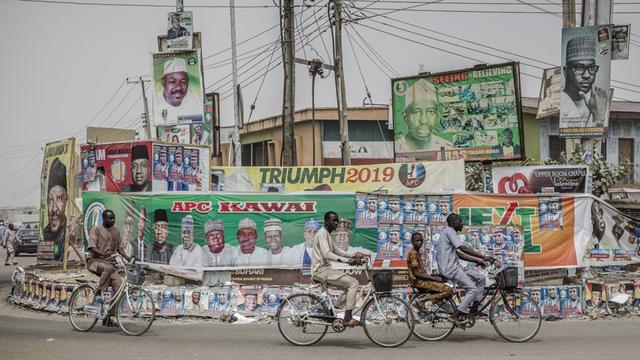 Menschen fahren auf Räder in der nigerianischen Stadt Mubi im Staat Adamawa an Wahlkampf-Plakaten vorbei. Die Wahl wird sich vor allem zwischen Präsident Muhammadu Buhari und seinem wichtigsten Herausforderer, Atiku Abubakar, entscheiden.