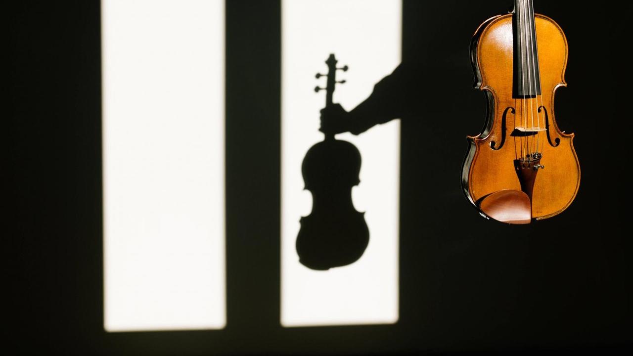 Eine Violine, die im Vordergrund im Licht zu sehen ist, wirft im Hintergrund einen markanten Schatten auf eine weiße Wand. Eine Violine, die im Vordergrund im Licht zu sehen ist, wirft im Hintergrund einen markanten Schatten auf eine weiße Wand.