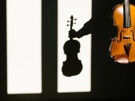 Eine Violine, die im Vordergrund im Licht zu sehen ist, wirft im Hintergrund einen markanten Schatten auf eine weiße Wand.