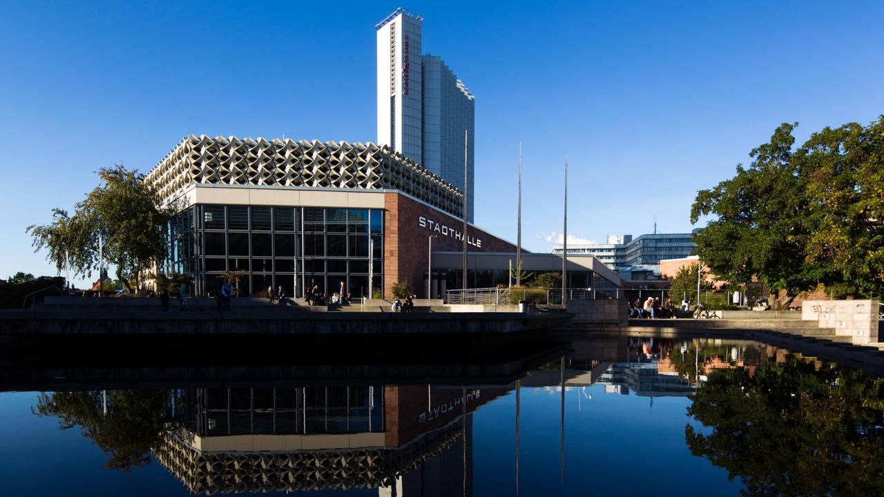 Ein moderner Gebäudekomplet von 1974 mit Flachbau und Hochhaus spiegelt sich in einer davor liegenden Brunnenanlage an einem Tag mit blauem Himmel. Ein moderner Gebäudekomplet von 1974 mit Flachbau und Hochhaus spiegelt sich in einer davor liegenden Brunnenanlage an einem Tag mit blauem Himmel.