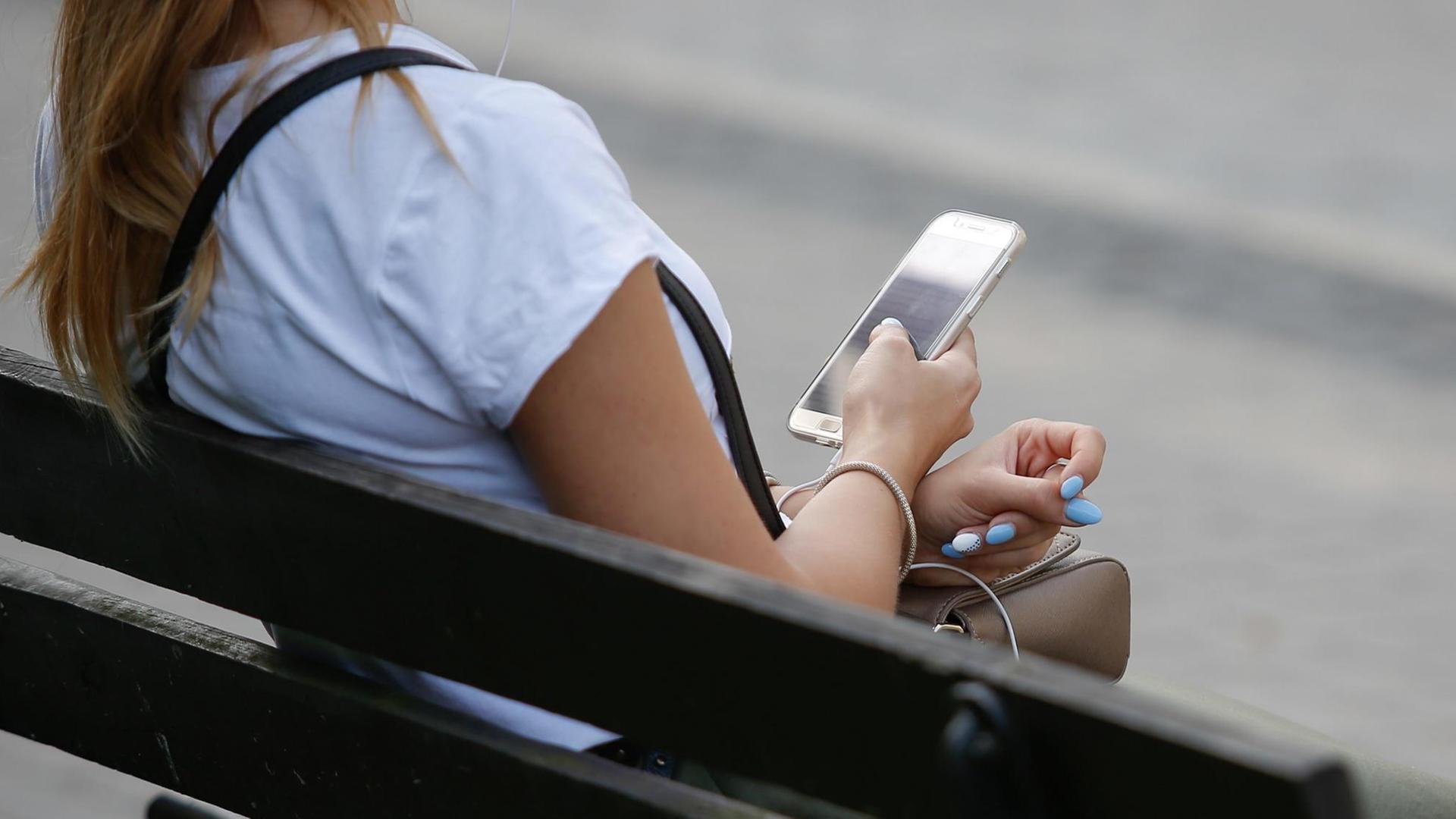 Eine Junge Frau sitzt auf einer Parkbank und blickt auf ihr Smartphone. Eine Junge Frau sitzt auf einer Parkbank und blickt auf ihr Smartphone.