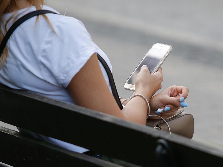 Eine Junge Frau sitzt auf einer Parkbank und blickt auf ihr Smartphone.